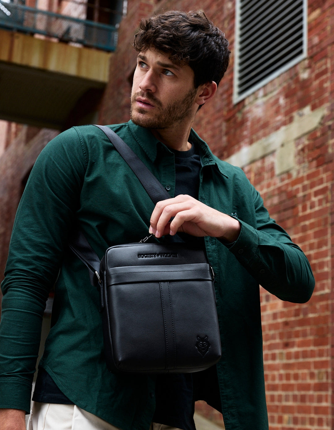 Man in a dark green shirt wearing a black leather cross body bag against a brick wall