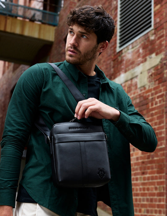 Man in a dark green shirt wearing a black leather cross body bag against a brick wall