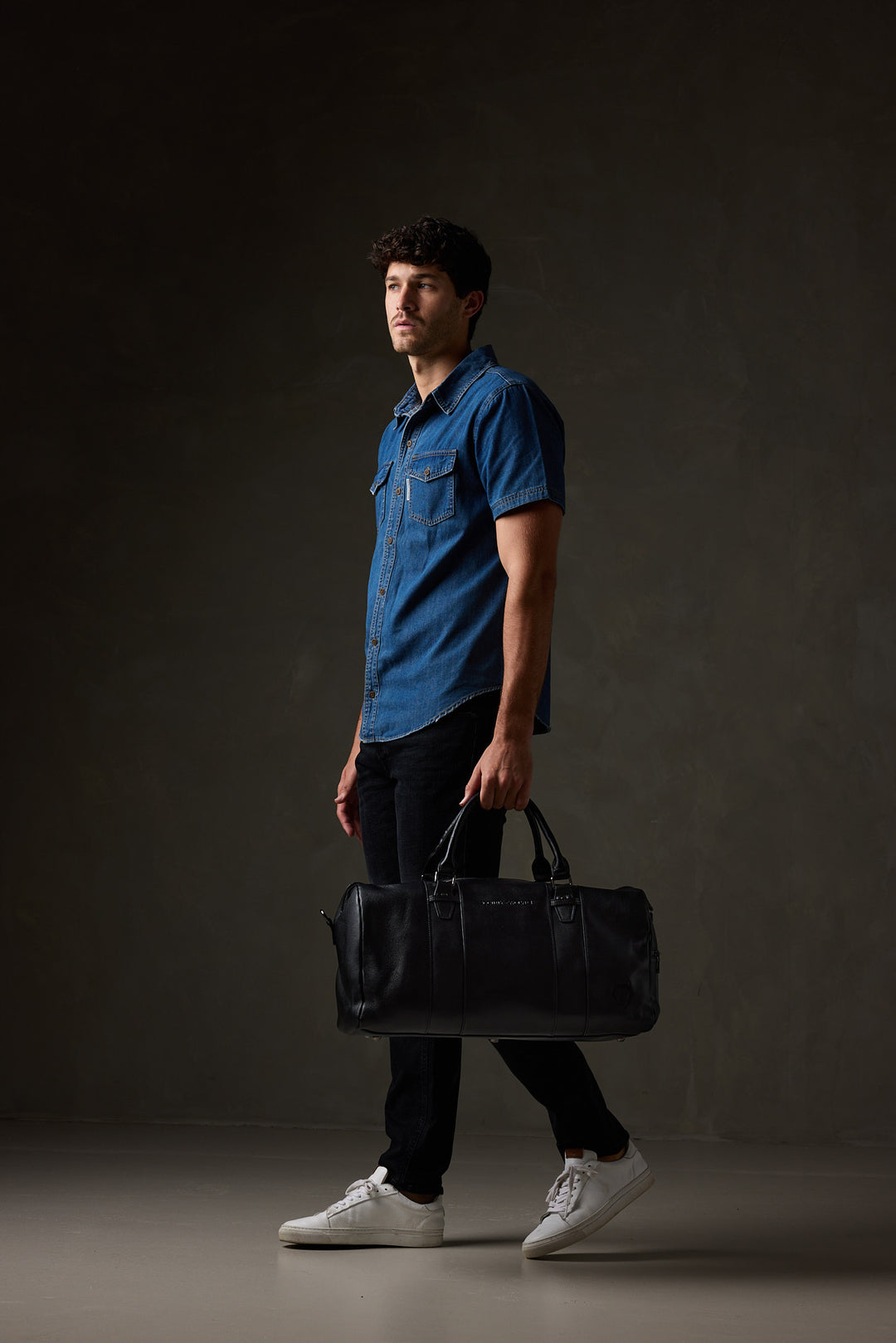 Man in a blue short-sleeve shirt, black trousers and white sneakers carrying a black leather overnight bag