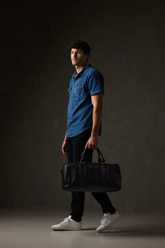 Man in a blue short-sleeve shirt, black trousers and white sneakers carrying a black leather overnight bag