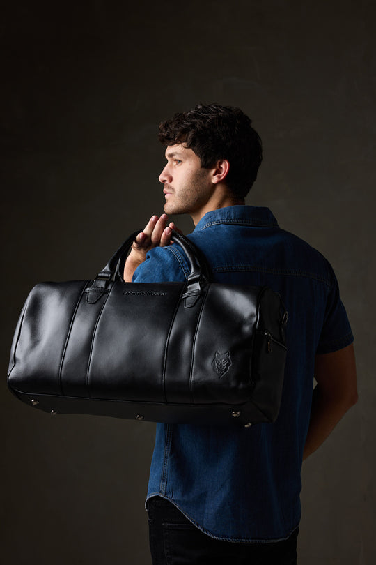 Man in a blue denim shirt carrying a black leather overnight bag over his shoulder against a dark studio background