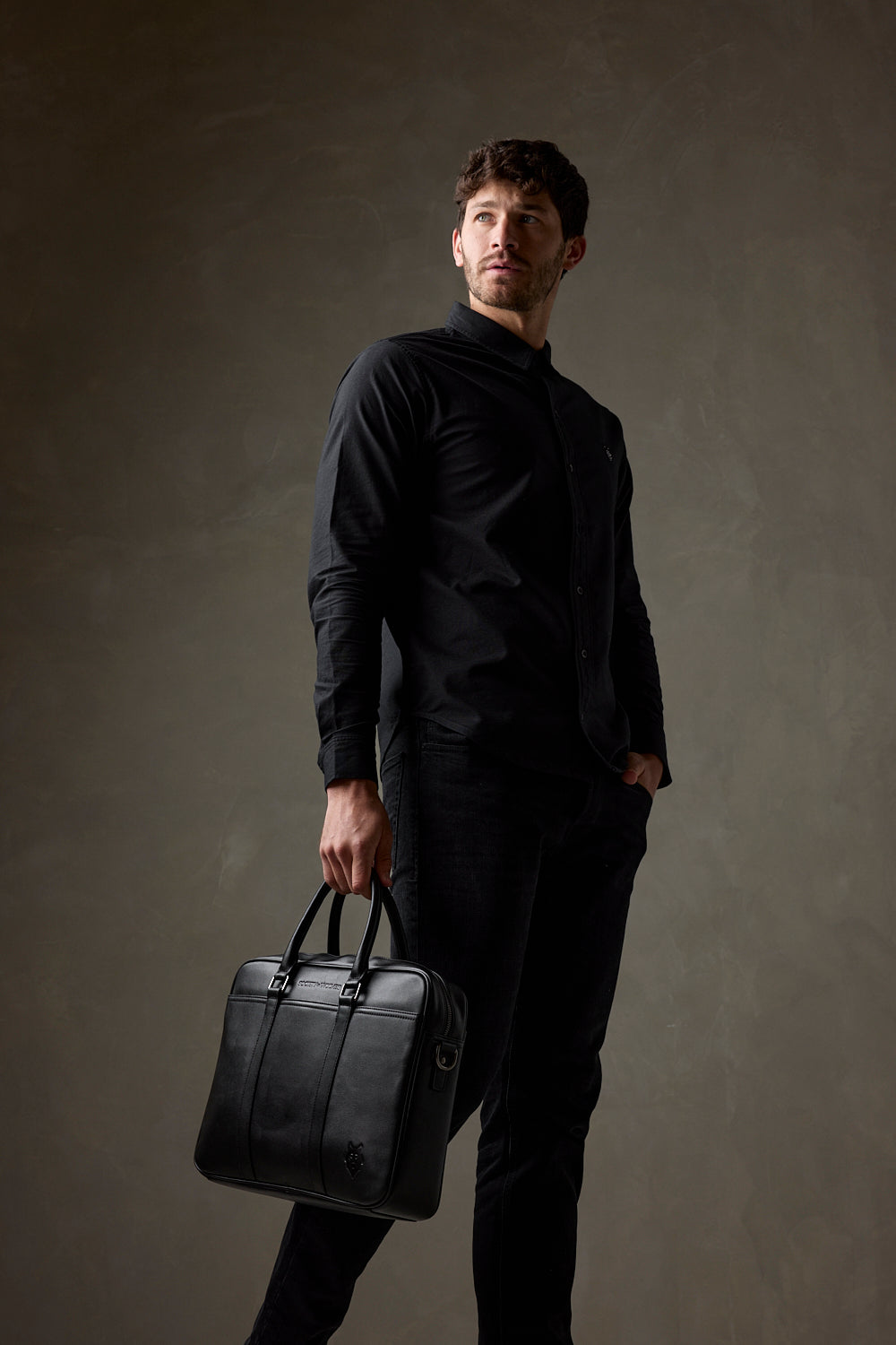 Man in all black carrying a black leather laptop bag by the handles against a dark studio background