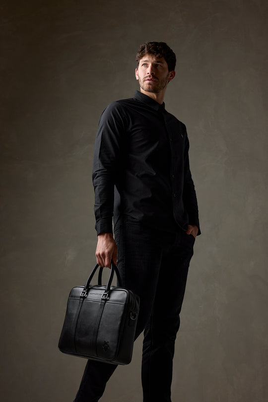 Man in all black carrying a black leather laptop bag by the handles against a dark studio background