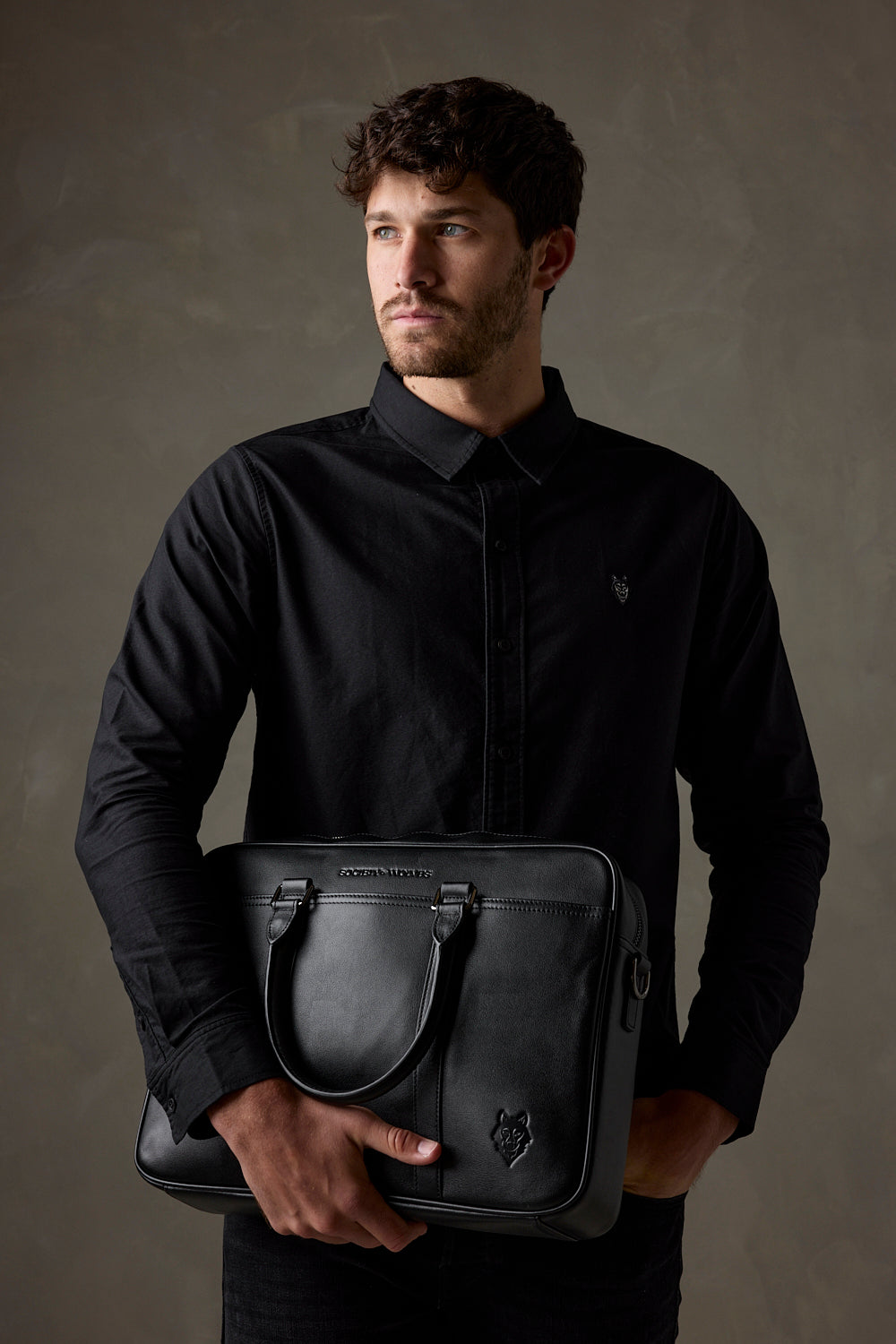 Man in a black shirt holding a black leather laptop bag against a dark studio background