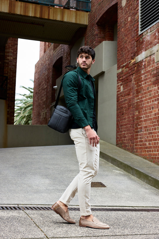 Man walking between brick buildings in a dark green shirt and off-white trousers with a black leather cross body bag
